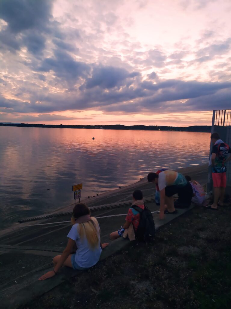 Podopieczni podczas wyjazdu wakacyjnego podziwiają zachód słońca nad jeziorem.